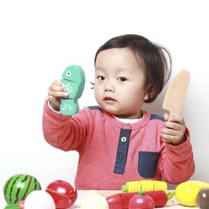 Photograph of a toddler, holding a toy dinosaur and wooden spatula