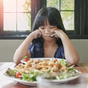Photograph of a young girl rubbing her eyes at a table with a plate of shrimp salad