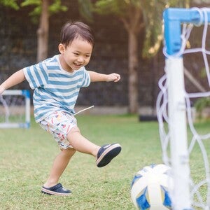 Photograph of a smiling young boy kicking a soccer ball toward a backyard goal on grass