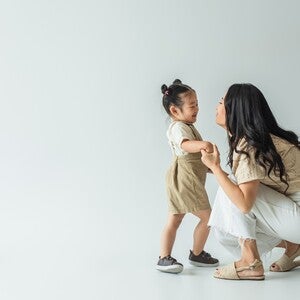 Photograph: woman kneeling, holding toddler's hands as they smile on white background