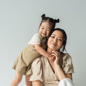 Photo: seated woman with child hugging her shoulders, neutral clothing, light gray background