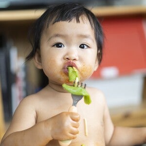 Toddler with blurred face eating green food, messy chest, holding spoon, colorful shelf behind