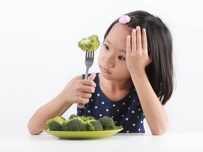 Photograph of a young girl holding broccoli on a fork, plate of broccoli in front