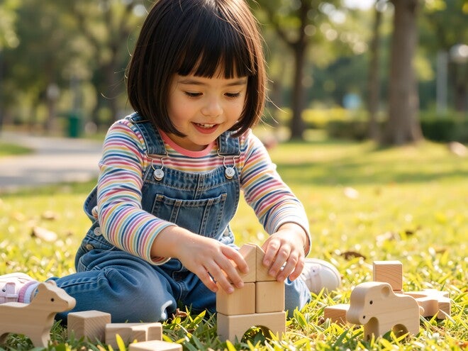 Photograph: smiling toddler in overalls arranging wooden blocks on grass in sunlit park