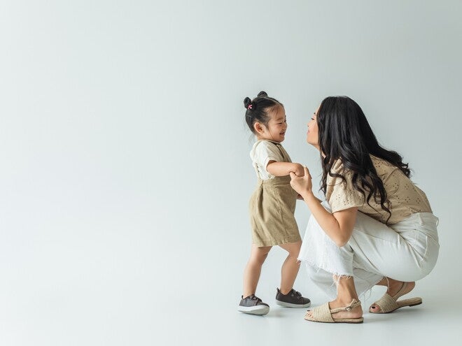 Photograph: woman kneeling, holding toddler's hands as they smile on white background