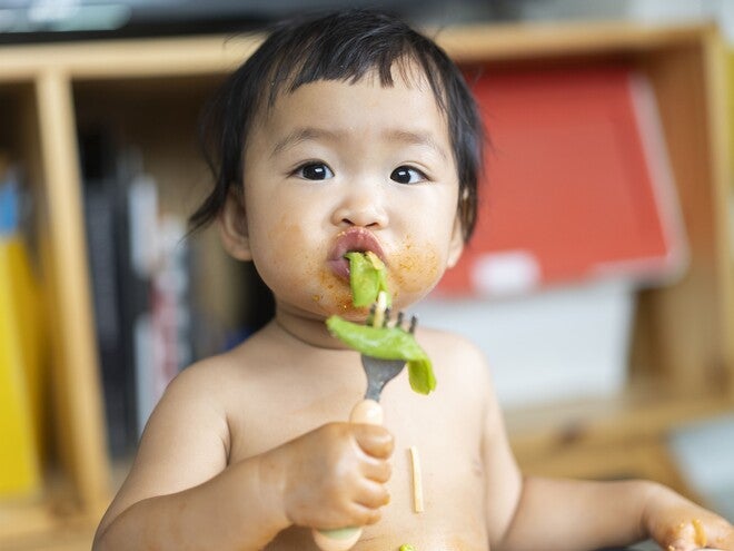 Toddler with blurred face eating green food, messy chest, holding spoon, colorful shelf behind