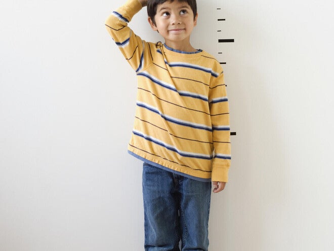 Photograph — smiling boy in yellow striped shirt by height chart, hand on head